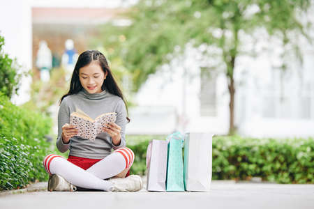 Pretty smiling teenage girl sitting on pavement next to shopping bags and reading a bookの写真素材