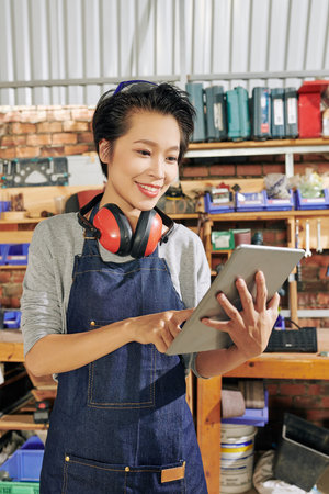 Pretty smiling young female carpenter with digital tablet checking dimensions of furniture item she...