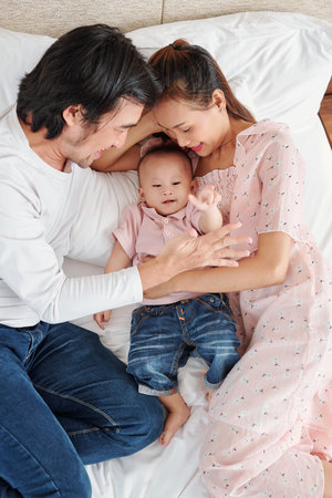 Positive young father and mother playing with little baby boy when lying on bed, view from above