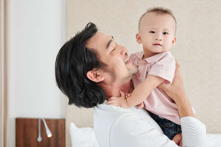 Joyful young Asian man carrying his little newborn son and looking at him with love and tenderの写真素材