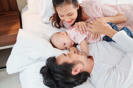 Young parents hugging their baby boy almost sleeping between them on comfy soft bedの写真素材