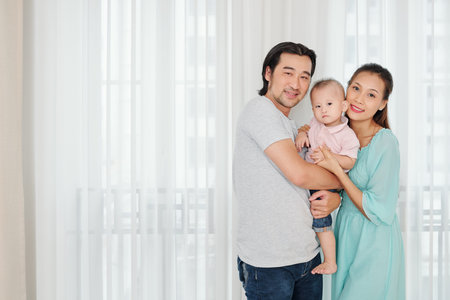 Happy young parents holding their serious little son and looking at camera