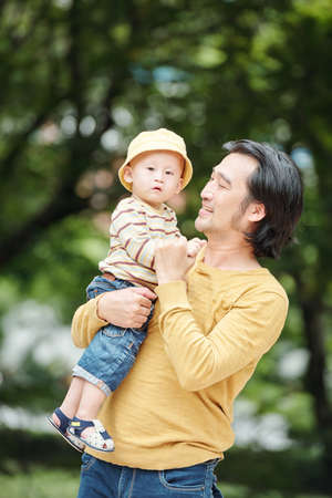 Portrait of positive young Vietnamese man carrying little son when walking in city parkの写真素材