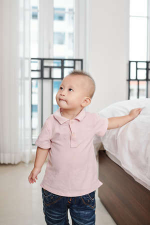 Curious cute little boy standing at bed, touching blanket and looking upの写真素材