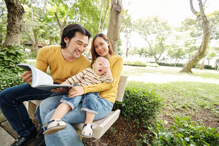 Adorable little kid sitting on laps of his mother and yawning as he is bored of his father reading a...