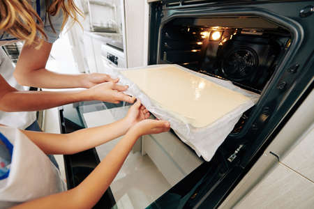 Close-up image of mother and daughter putting baking sheet in hot oven when making sponge cake at homeの写真素材