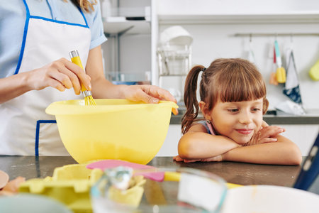 Smiling little girl leaning on kitchen counter and watching cartoon on tablet computer when her...