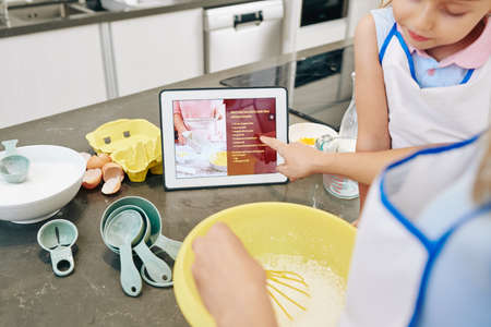 Woman pointing at recipe on screen of tablet computer when baking cake for her daughterの写真素材