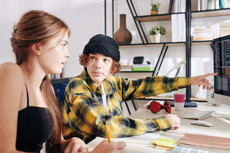 Teenage boy in plaid shirt and hat pointing at computer screen with programming code when doing homework with his sister or friend at homeの写真素材