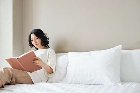 Pretty Asian woman sitting on bed and reading interesting bookの写真素材