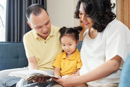 Smiling Vietnamese mother and father showing interesting book to little daughetr