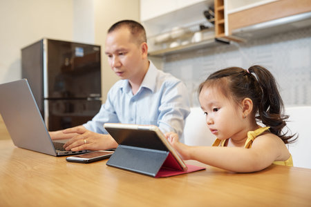 Serious Vietnamese man answering e-mails on laptop when daughter sitting next to him and using...