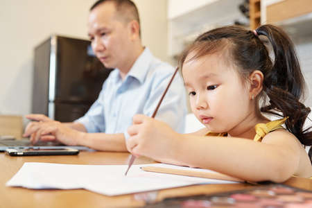Little Vietnamese girl enjoying drawing with pencil when sitting at table next to her father busy with work on laptopの写真素材