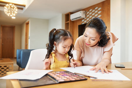 Pretty Asian woman helping daughter with painting when they are spending time at home due to...