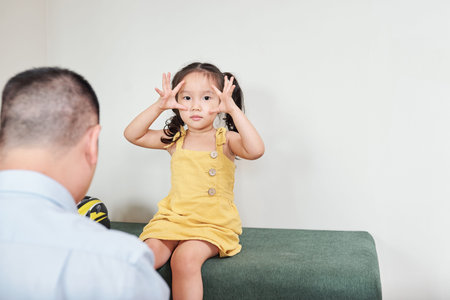 Little Vietnamese girl sitting on soft bench and making wide eyes gesture when father putting on her...