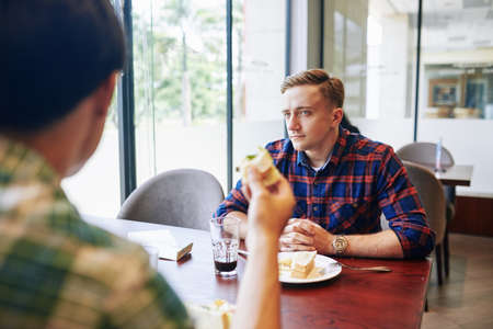 Serious handsome young man and plaid shirt sitting at cafe table and listening to story of his friendの写真素材