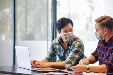 Positive young people in medical masks sitting at office table and discussing way of online business...