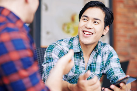 Happy smiling Asian young man with smartpone in hands talking to his friend when they are sitting at...