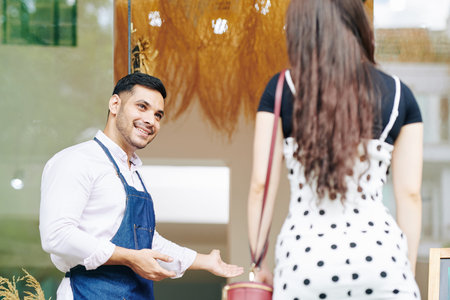 Positive handsome young small cafe owner making welcoming gesture and inviting female customer to...