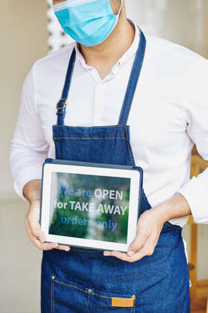 Waiter in medical mask showing tablet computer that says that they are open to take away orders only due to pandemicの写真素材