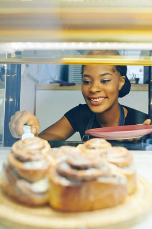 Happy pretty smiling Black waitress taking cinnamon bun from showcase to serve for customersの写真素材