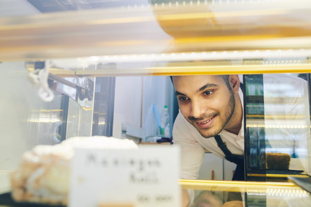 Smiling handsome barista taking piece of tasty cake from shop window for customer