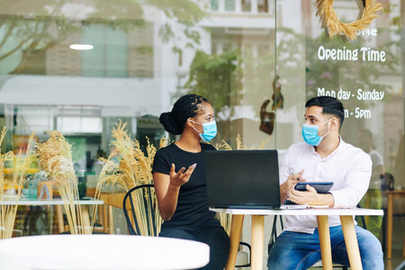 Multi-ethnic entrepreneurs in medical masks meeting at cafe table and discussing business strategy