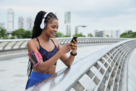 Smiling attractive young mixed-race woman standing on bridge and checking track of her run via...
