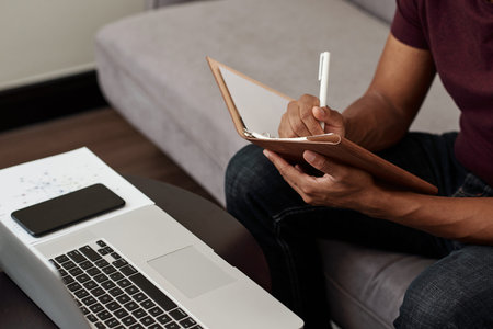 Close-up image of university student watching online webinar on laptop and taking notes in copybook