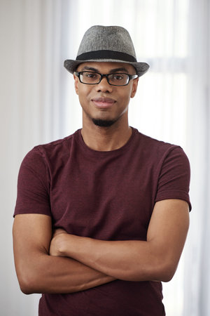 Portrait of handsome smiling young Black man in fedora hat and glasses crossing hands and smiling at...