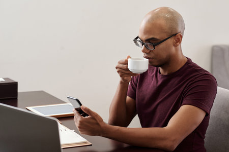 Young Back African-American man in glasses drinking coffee and checking messages and notifications...