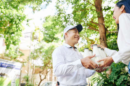 Happy smiling Vietnamese delivery man giving set of milk bottles to pretty young womanの写真素材
