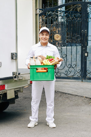 Portrait of handsome young Vietnamese courier holding plastic crate with milk bottles, fresh bread...