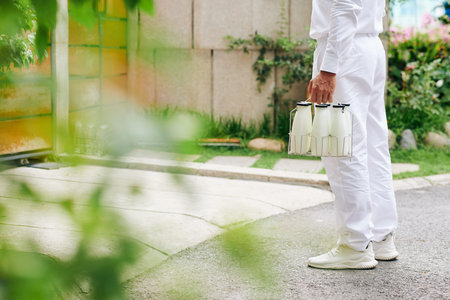 Cropped image of milkman in white uniform standing at house entrance with glass bottles of milk or...