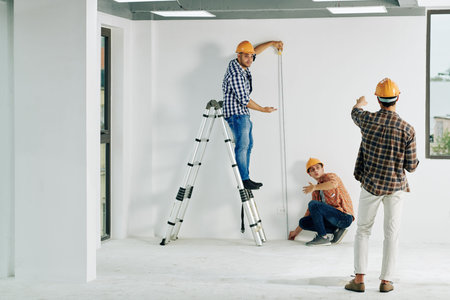Team of construction workers in hardhats measuring wall with tape measure
