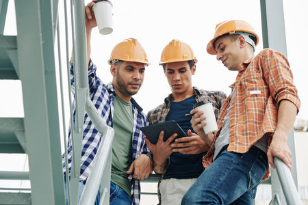 Young construction workers in hardhats drinking take away coffee and checking e-mail from contractor...
