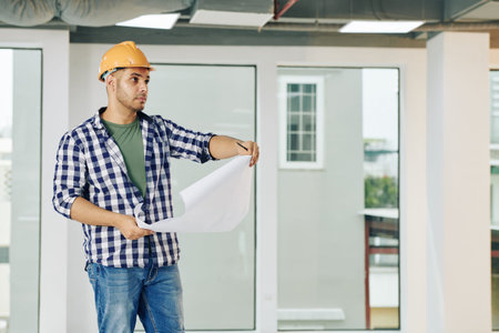 Serious handsome young construction engineer with blueprint checking service lines in building