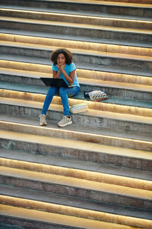 Curly teenage scoolgirl sitting on steps outdoors, reading book and writing essay on laptop for...