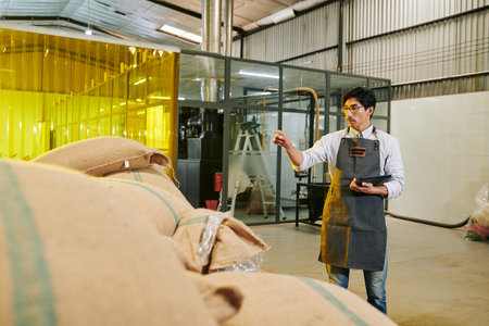 Roastery owner with digital tablet in hands counting sacks with coffee beans in warehouse