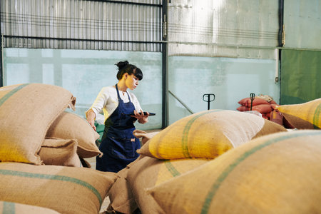 Serious female roastery worker with digital tablet in hands walking long sacks of coffee beans in...