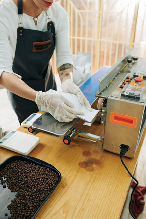 Female roastery worker in apron and rubber gloves sealing seams of coffee packages