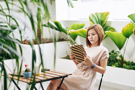 Serious young Asian woman reading captivating book at cafe table
