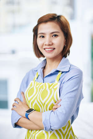 Portrait of pretty Vietnamese housewife in apron folding arms and smiling at cameraの写真素材