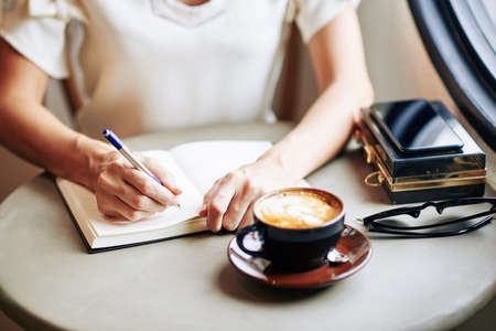 Close-up image of woman drinking cup of cappuccino and writing thoughts and ideas in gratitude journalの写真素材