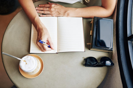 Hands of young woman drinking cappuccino and writing plans for the day in planner, view from above