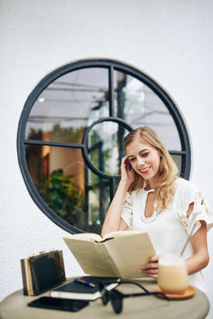 Beautiful smiling young blond woman reading funny story in thick book when resting at outdoor cafeの写真素材