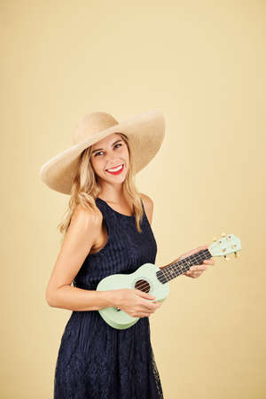 Portrait of smiling pretty blond young woman in straw hat playing ukulele, isolated on yellowの写真素材