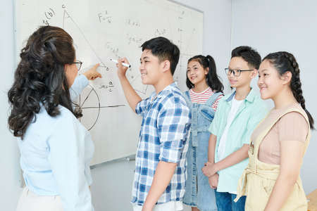 Science teacher explaining theorem to group of students and asking schoolboy to writing equation on whiteboardの写真素材