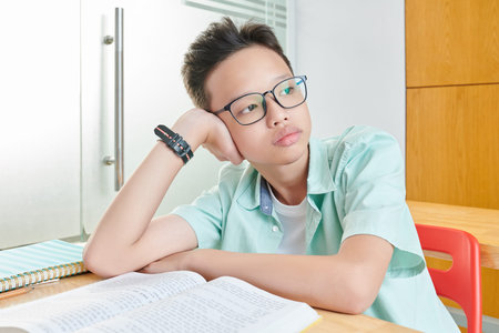 Portrait of pensive bored Vietnamese schoolboy in glasses leaning on hand and looking through window