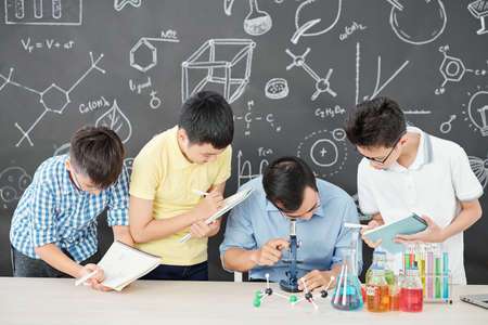 Science teacher looking through microscope at bacteria in petri dish when school students standing near by and writing in textbooksの写真素材
